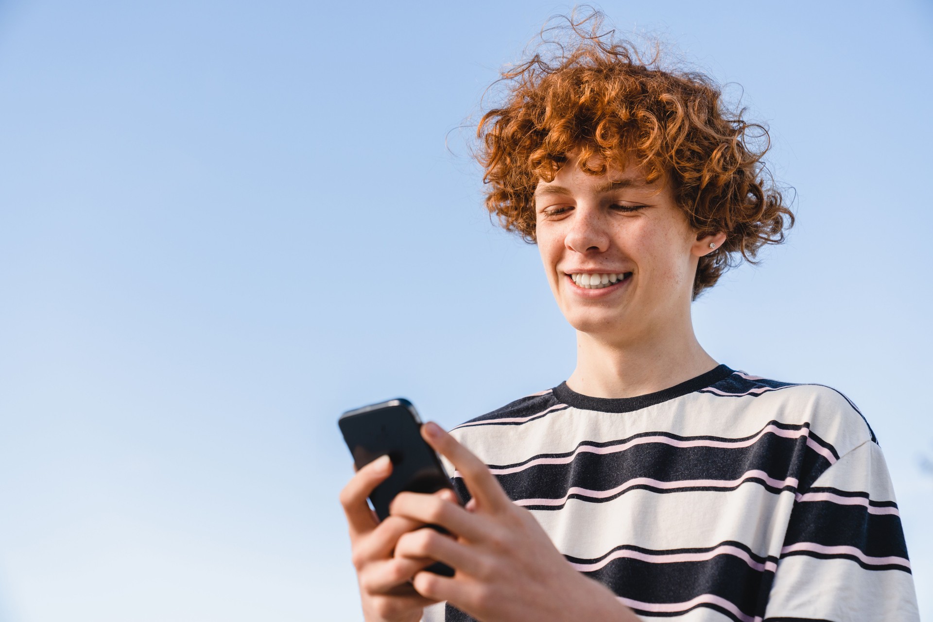 Smiling teen boy in casual holding using smart phone texting chatting with friends standing outdoor
